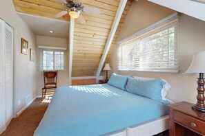 Un dormitorio con techo inclinado, una cama con ropa de cama azul, una mesita de noche con lámpara y una ventana con persianas.