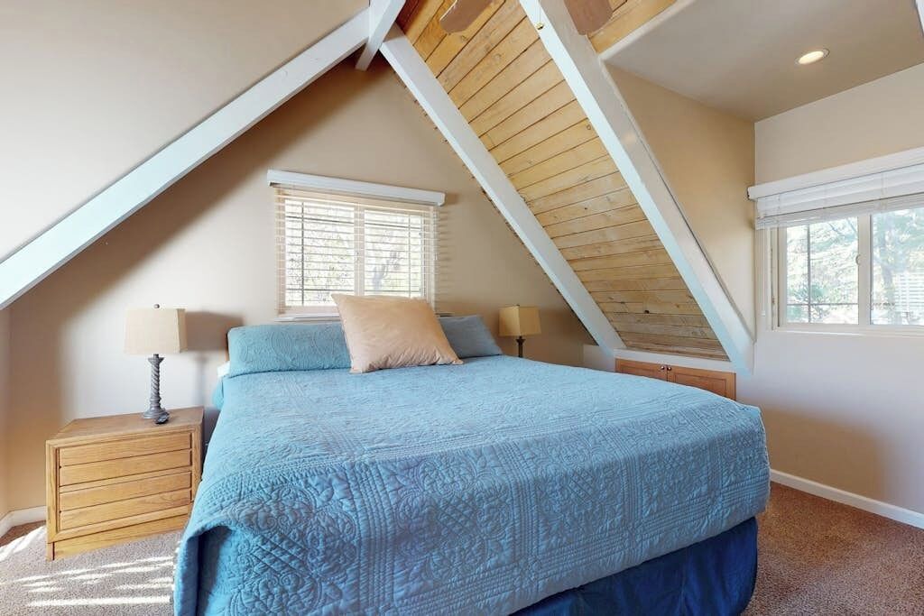 Quarto com teto inclinado, uma cama com colcha azul, duas mesas de cabeceira com luminárias e uma janela com persianas.