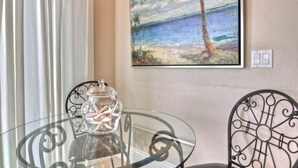 Een glazen tafel met een decoratieve pot, een schilderij van een strandtafereel en metalen stoelen.