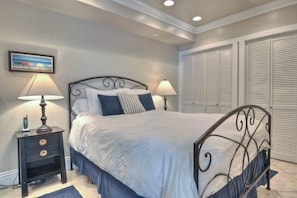 A bedroom with a bed, two bedside tables with lamps, a painting on the wall, and a set of white louvered closets.