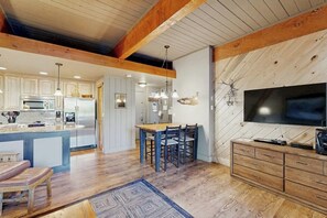 Eine moderne Küche mit Holzbalken, einem Essbereich mit Stühlen, einem an einer Holzwand montierten Fernseher und einem eingebauten Holzkasten.