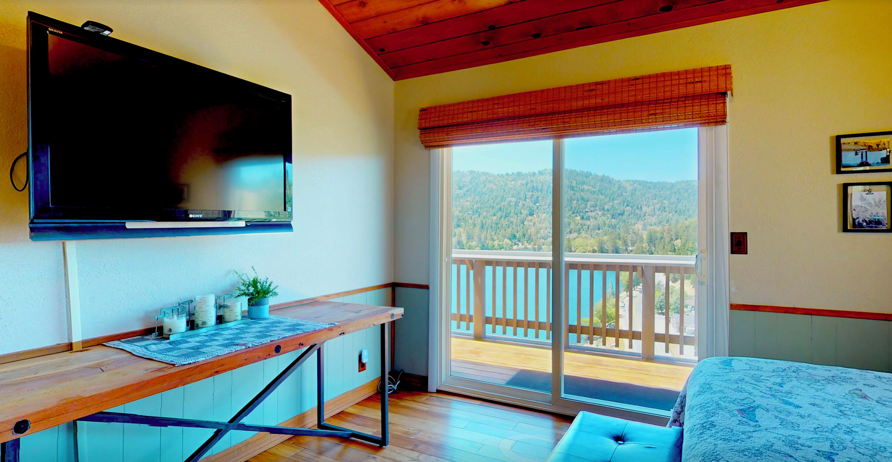 A room with a television, a desk with a plant, a bed, and a balcony with a view of the mountains.