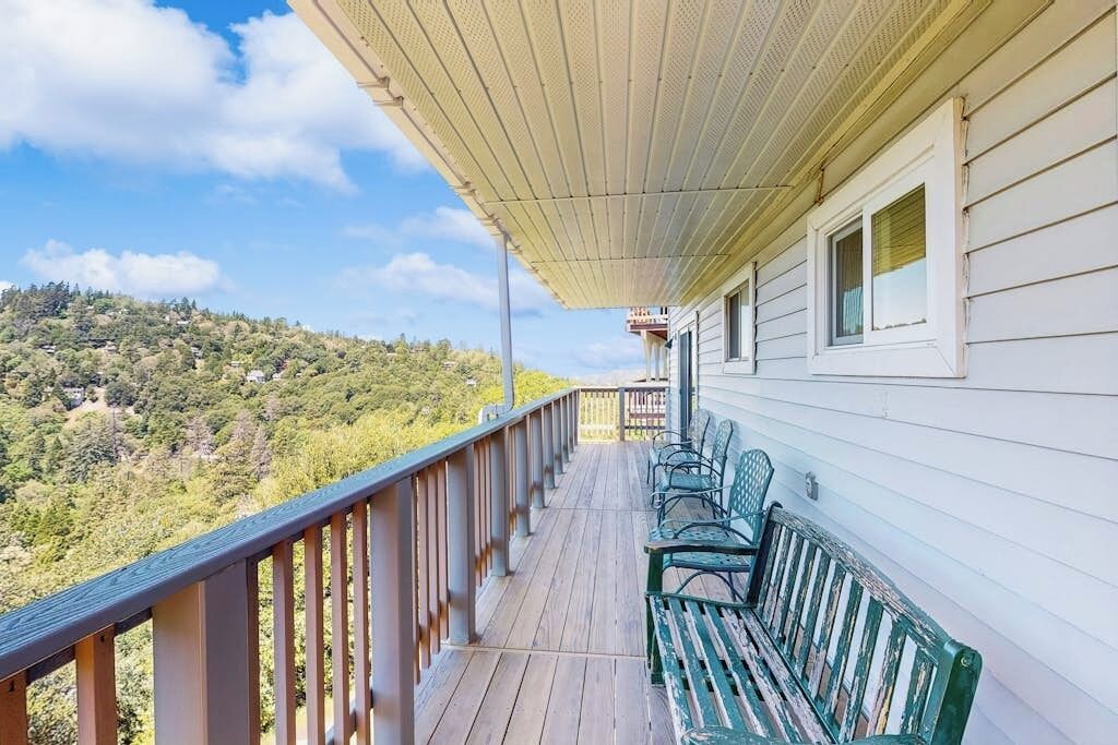 Une véranda couverte avec une balustrade en bois, un banc et une vue sur une colline boisée.