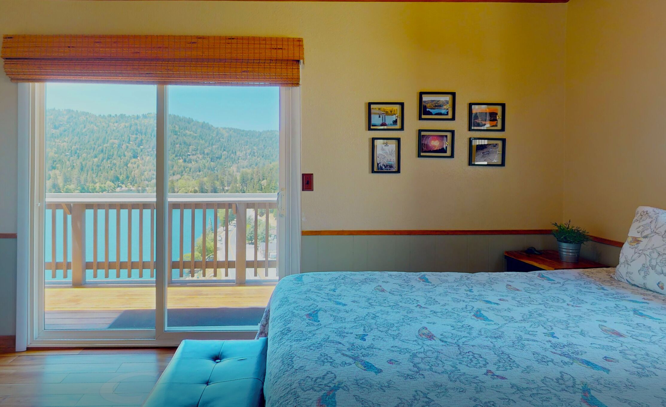 Une chambre avec un grand lit, un balcon donnant sur une zone boisée et plusieurs tableaux encadrés accrochés au mur.