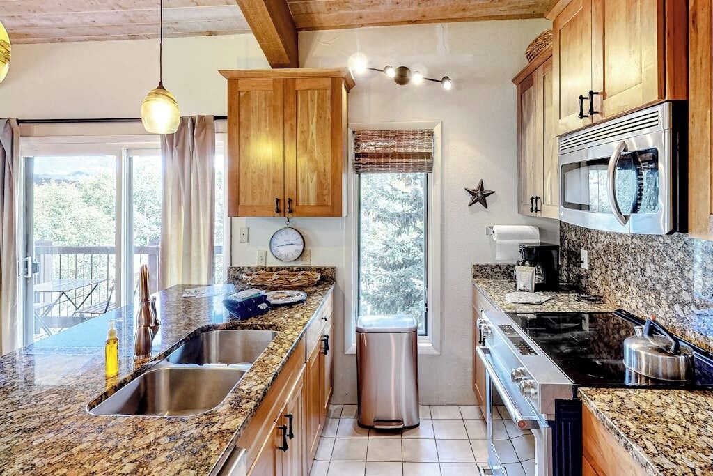 A kitchen with granite countertops, wooden cabinets, stainless steel appliances, and a window with a view of trees.