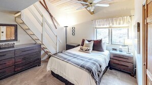 A bedroom with a bed, a dresser, a nightstand, a ceiling fan, a window with curtains, and a staircase. - Spacious condo with fireplace & balcony (Snowmass Village)