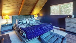 Een slaapkamer met een schuine kap, een bed met beddengoed met een patroon, een nachtkastje met een lamp, een dressoir en een raam met jaloezieën.