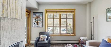 Een woonkamer met een open haard, een groot raam met uitzicht, een stoel, een bank en een poster aan de muur.