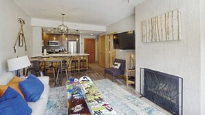 Een moderne woonkamer met een bank, een salontafel met een skateboard, een televisie, een open haard en een keuken op de achtergrond.