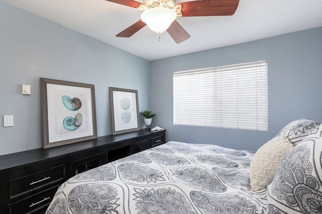 Une chambre à coucher avec un lit, un ventilateur de plafond, des œuvres d’art encadrées et une fenêtre avec des stores.