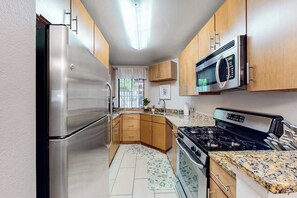 A compact kitchen with a stainless steel refrigerator, a microwave, a stove, wooden cabinets, and a granite countertop. - Pacific Cove | Pool & Hot Tub, Fitness Center | Walk to Beach & Dining (Playa Del Rey)