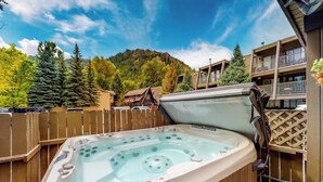 Un bain à remous installé à l’extérieur, entouré d’une clôture en bois, avec une montagne en arrière-plan et un bâtiment doté de balcons.