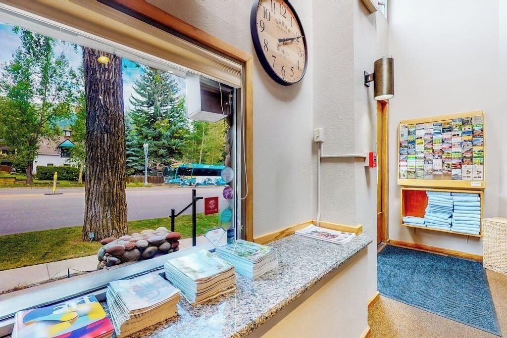 Un mostrador de recepción con un reloj, vista por la ventana a un árbol y una carretera, y un tablón de anuncios con folletos.