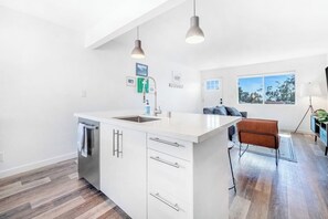 A modern kitchen with white cabinetry, a central island, pendant lighting, and a view to a living area with a sofa and a window. - Dog-friendly duplex with washer/dryer, two full kitchens, & large backyard (San Clemente)