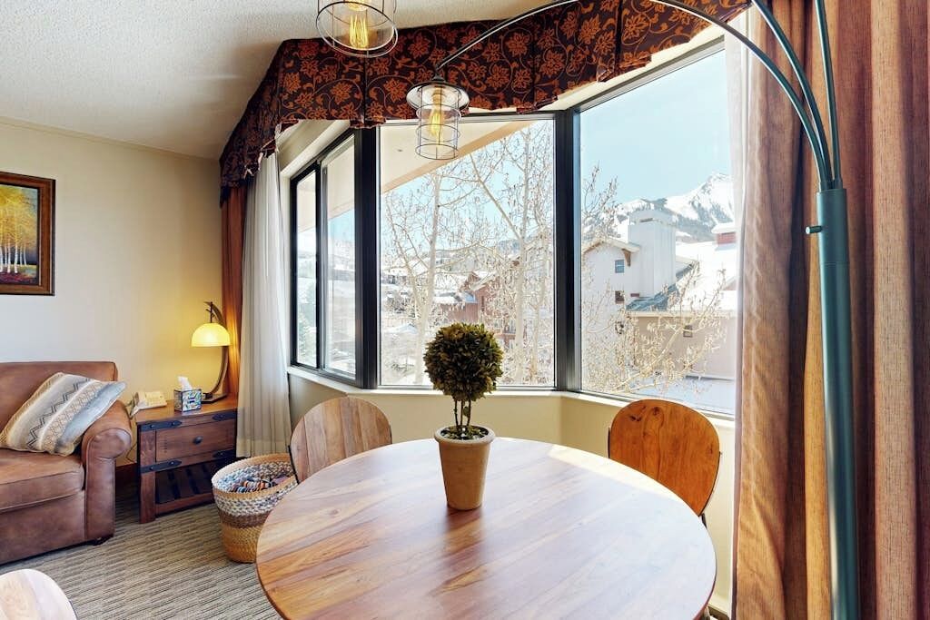 Une pièce chaleureuse avec une table ronde en bois, un canapé en cuir marron, une petite plante en pot et une fenêtre donnant sur des bâtiments et des arbres.
