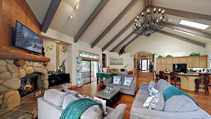A spacious living room with a stone fireplace, a large gray sofa, and a chandelier. - Beautiful home W/ Lake Access, Decks, Fire pit, Papoose Lake views, & EV Charger (Lake Arrowhead)