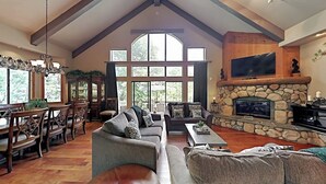 A spacious living room with a stone fireplace, a large TV, and a dining area with wooden furniture. - Beautiful home W/ Lake Access, Decks, Fire pit, Papoose Lake views, & EV Charger (Lake Arrowhead)