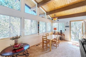 A spacious room with large windows, a dining area with wooden chairs, a black leather sofa, and a chandelier.