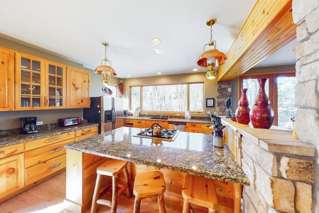 A kitchen with granite countertops, wooden cabinets, a central island with stools, and a stone fireplace.