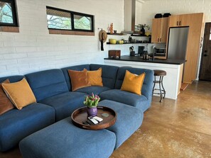 Ein modernes Wohnzimmer mit einem blauen, modularen Sofa, einem Holztisch und einer Küchenzeile mit Holzausziehtüren und einem Edelstahlkühlschrank.