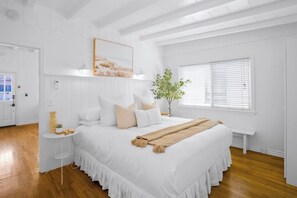 Ein Schlafzimmer mit einem großen Bett, weißen Wänden, Holzboden, einem Nachttisch mit Lampe und einem Fenster mit Jalousien.