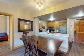 A dining area with a wooden table and chairs, a kitchen with modern appliances, and a red couch in the background. - Walk to slopes - Village condo with balcony, views, hot tubs, & gym (Copper Mountain)