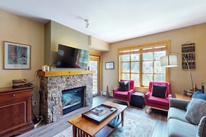 A living room with a stone fireplace, a flat-screen TV, a red armchair, a grey sofa, and a large window with a view of trees. - Walk to slopes - Village condo with balcony, views, hot tubs, & gym (Copper Mountain)