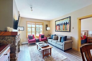 A living room with a stone fireplace, a large window, a colorful wall mural, and a cozy seating area. - Walk to slopes - Village condo with balcony, views, hot tubs, & gym (Copper Mountain)