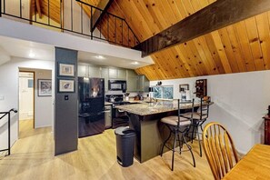 A modern kitchen with a central island, a dining area with chairs, and a staircase leading to an upper level. - Enchanting lake retreat with fireplace, games, & great deck views - dog-friendly (Crestline)