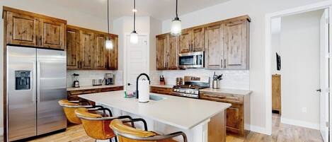 A modern kitchen with wooden cabinets, stainless steel appliances, a central island with bar stools, and pendant lighting.