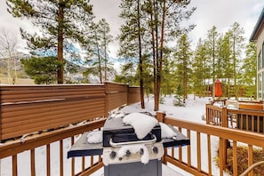 Eine Terrasse mit Grill, Holzgeländern und verschneiter Landschaft mit Bäumen.