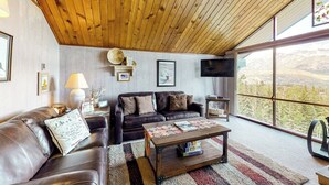 Una sala de estar con techo de madera, sofá de cuero, una mesa de centro y un ventanal con vistas a árboles.