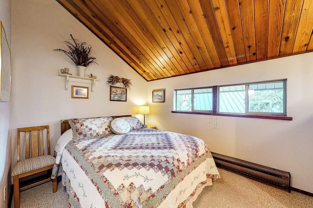 A bedroom with a wooden ceiling, a bed with a patterned quilt, a chair, a radiator, and a window with a view.
