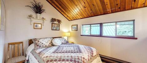 A bedroom with a wooden ceiling, a bed with a patterned quilt, a chair, a radiator, and a window with a view.