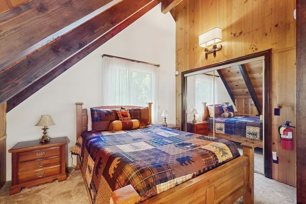 Quarto com teto de madeira, um espelho grande, uma cama com colcha, uma mesa de cabeceira e um abajur.