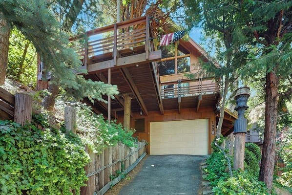 A two-story wooden cabin with a garage, surrounded by trees and a wooden fence. - Rustic, wooded escape near the village w/ a lovely deck (Lake Arrowhead)