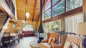A cozy cabin interior with a wooden ceiling, large windows, a fireplace, a dining area, and a sofa. - Rustic, wooded escape near the village w/ a lovely deck (Lake Arrowhead)