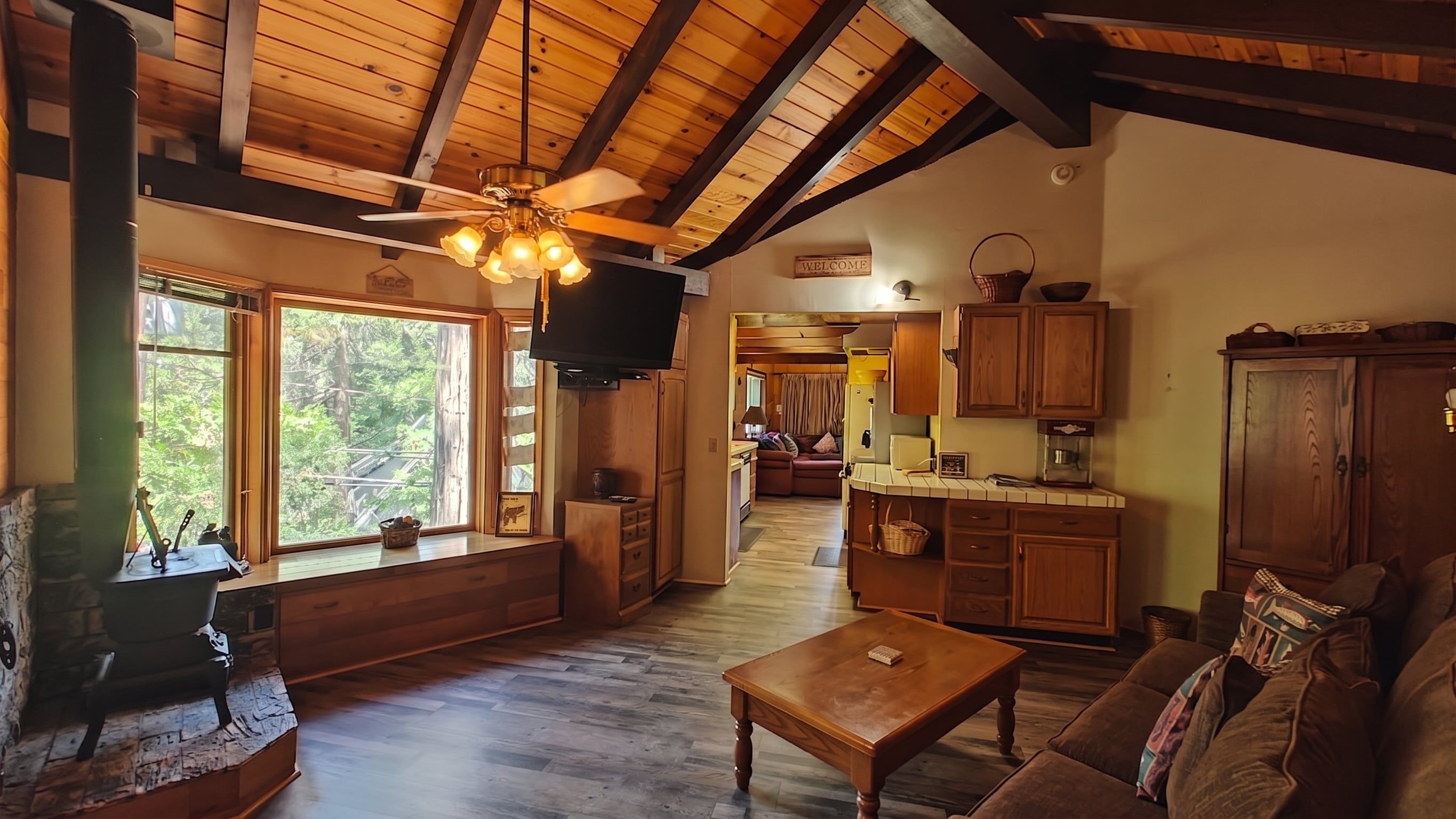 A cozy cabin interior with a wooden ceiling, a television mounted on the wall, a sofa, a coffee table, and a kitchen area with wooden cabinets.