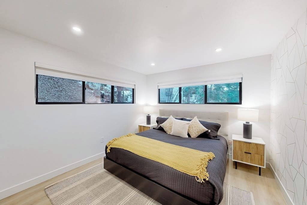 A modern bedroom with a large bed, two bedside tables, and a view of trees through the windows.