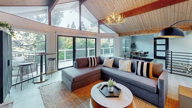 A modern living room with a sectional sofa, a coffee table, and a dining area with a table and chairs. - Dog-friendly home with beautiful views, large deck, smart TVs & lake access (Lake Arrowhead)