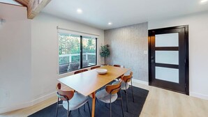 A modern dining area with a wooden table and chairs, a potted plant, and a large window. - Dog-friendly home with beautiful views, large deck, smart TVs & lake access (Lake Arrowhead)
