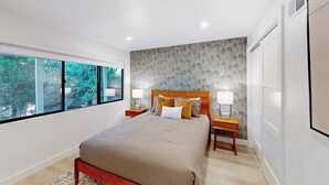 A modern bedroom with a large bed, nightstands, and a view of trees through large windows. - Dog-friendly home with beautiful views, large deck, smart TVs & lake access (Lake Arrowhead)