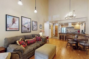 Ein Wohnzimmer mit einer Couch, Hocker und einem Essbereich mit Tisch und Stühlen. An den Wänden hängen Bilder und über dem Esstisch hängt eine Chandelier.