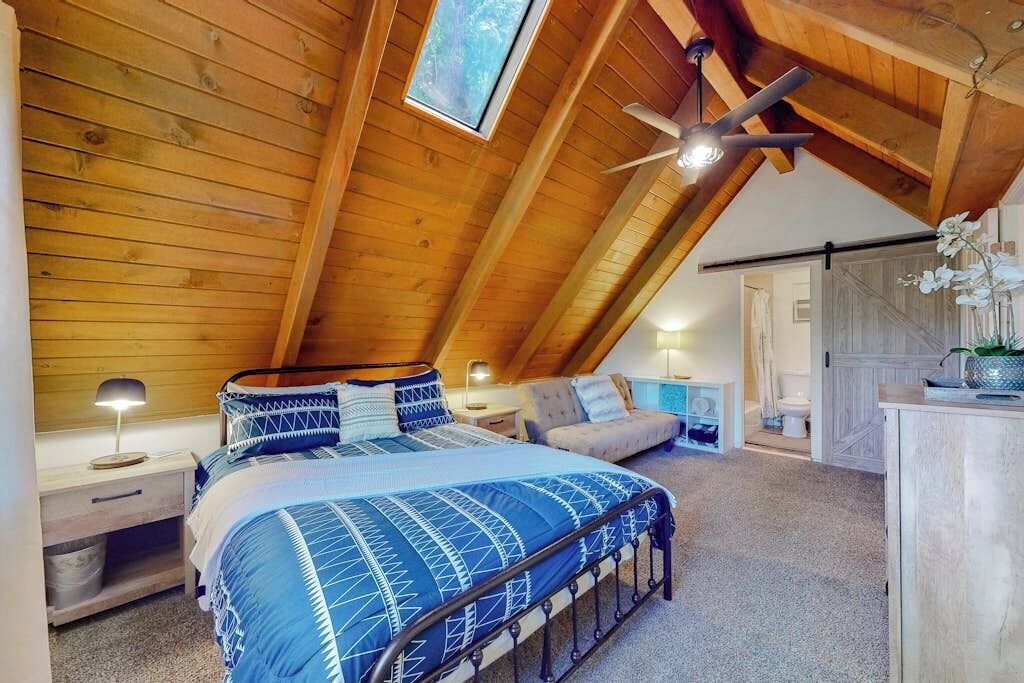 Une chambre avec un plafond en pente, un lit recouvert d’une couvre-lit à motifs bleus, une table de chevet, une lampe de chevet, un ventilateur de plafond et une fenêtre donnant sur les arbres.