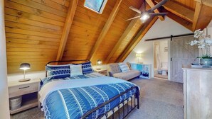 A bedroom with a sloped ceiling, a bed with a blue patterned bedspread, a nightstand, a bedside lamp, a ceiling fan, and a window with a view of trees. - Tree-surrounded retreat w/ balcony, patio, fireplace, Lake access, washer/dryer (Lake Arrowhead)
