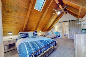 Ein Schlafzimmer mit schrägen Wänden, einem Bett mit blau gemustertem Überwurf, einem Nachttisch, einer Nachttischlampe, einem Deckenventilator und einem Fenster mit Blick auf Bäume.