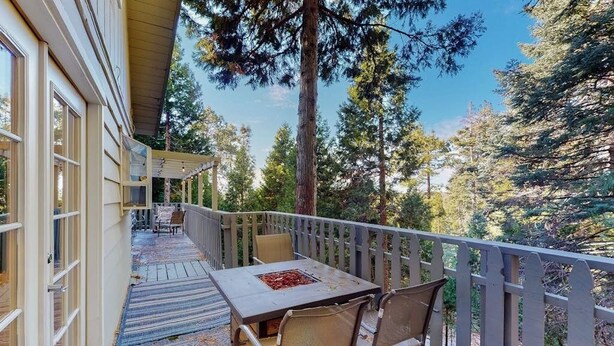 A covered outdoor area with a table and chairs, surrounded by a wooden railing, overlooking a forested landscape. - Tree-surrounded retreat w/ balcony, patio, fireplace, Lake access, washer/dryer (Lake Arrowhead)