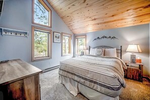 A bedroom with a sloped ceiling, a bed with a striped bedspread, two bedside lamps, a nightstand, and a window with a view of trees. - Delightful mountain retreat, NEW private hot tub, fireplace, deck, & great views (Durango)