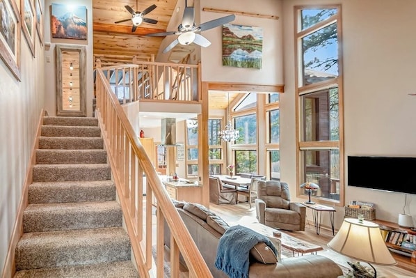 A spacious living area with a staircase, large windows, and a ceiling fan. - Delightful mountain retreat, NEW private hot tub, fireplace, deck, & great views (Durango)