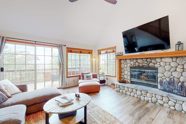 A spacious living room with a stone fireplace, a large flat-screen TV mounted above it, and a comfortable seating area with a sofa and a coffee table. - Spacious home with three decks, views, game room, fireplace, & fast WiFi (Lake Arrowhead)